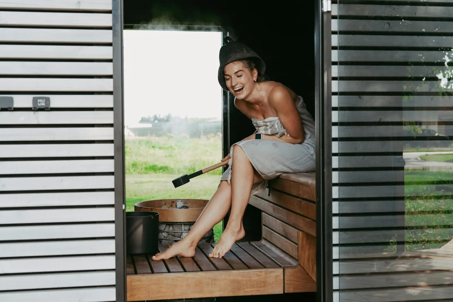 A woman in a felt sauna hat laughing as she pours water, steam rising, green field beyond the open doorway