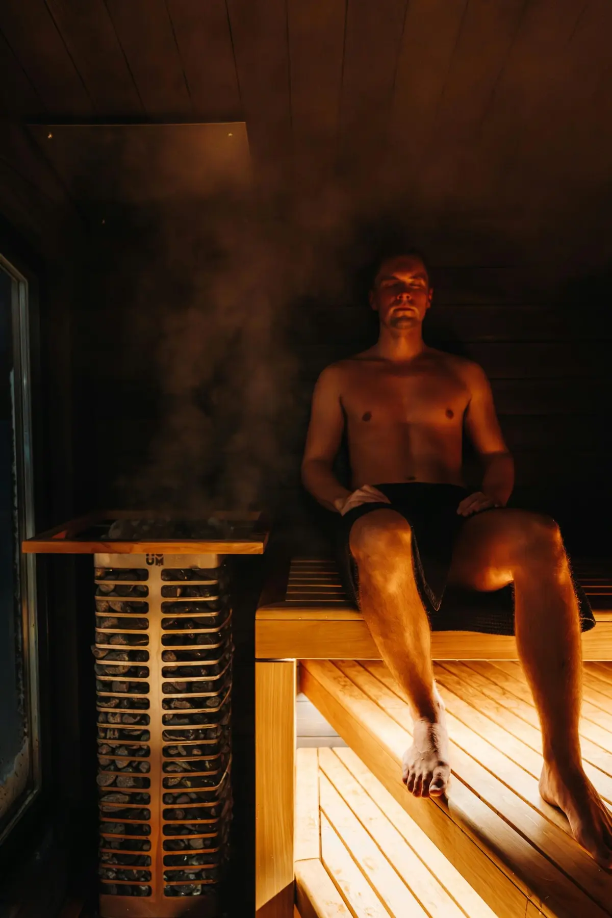 A man resting in amber light beside the glowing heater, steam rising off the stones